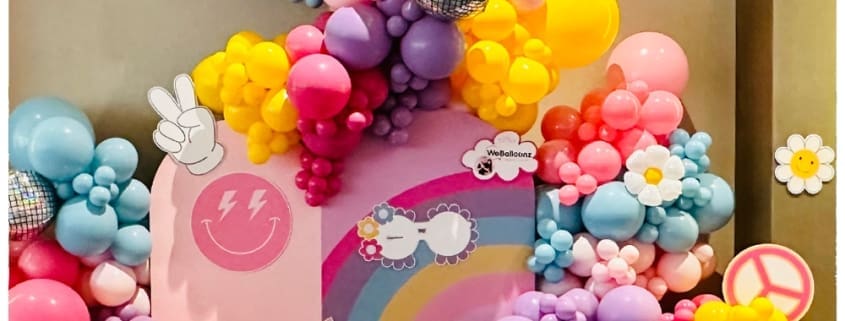 Purple, pink, blue, and yellow balloon display with balloon flowers, disco balls and 60s cut outs scattered in balloons.